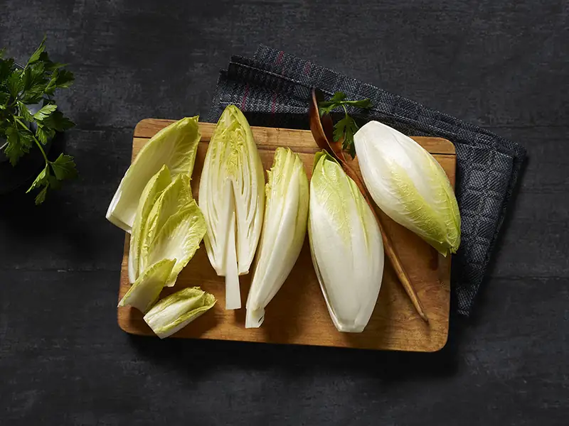 Mehrere Stück weißer Chicorée arrangiert auf einem Holzbrett, teilweise halbiert und Strunk herausgeschnitten