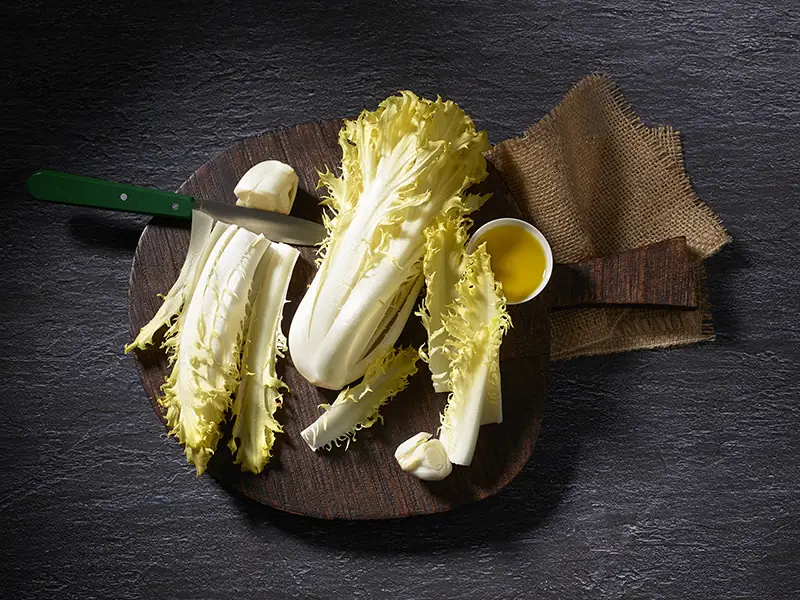 Friseline (gekräuselter Chicorée) auf einem Holzbrett, arrangiert mit einem Schälchen Olivenöl. ans sieht, dass man nur einen Zentimeter com Strunk wegschneiden muss, um den Salat vorzubereiten.